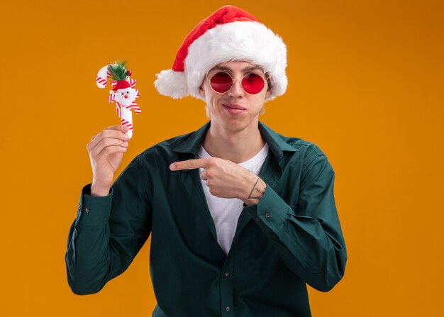 Heureux jeune homme blond portant bonnet de noel et lunettes tenant et pointant sur l'ornement de canne douce regardant la caméra isolée sur fond orange