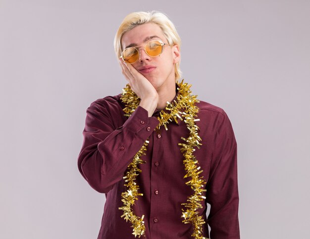 Heureux jeune homme blond portant des lunettes avec guirlande de guirlandes autour du cou en gardant la main sur le visage en regardant la caméra isolée sur fond blanc