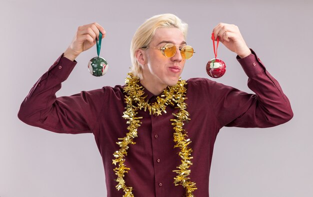 Heureux jeune homme blond portant des lunettes avec guirlande de guirlandes autour du cou tenant des boules de Noël près de la tête isolé sur mur blanc