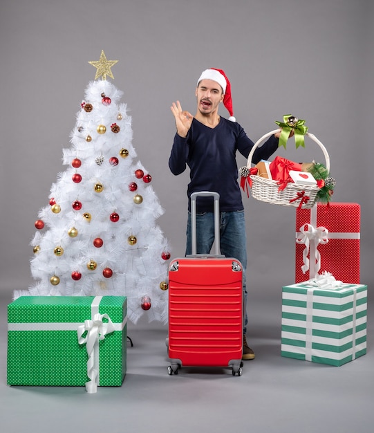 Heureux jeune homme avec bonnet de Noel tenant un panier-cadeau montrant signe okey près de l'arbre de Noël sur fond gris