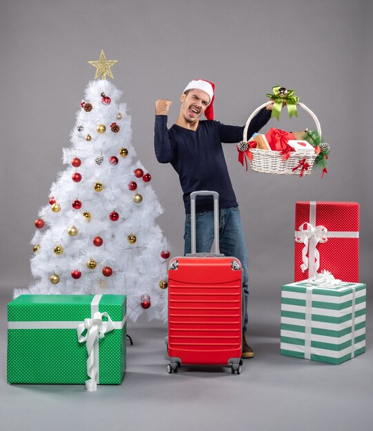 Heureux jeune homme avec bonnet de Noel tenant un panier-cadeau près de l'arbre de Noël sur fond gris