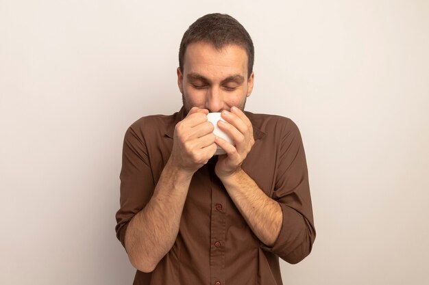 Heureux jeune homme caucasien, boire une tasse de thé avec les yeux fermés isolé sur fond blanc avec copie espace