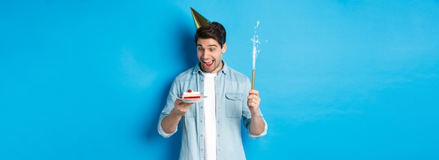 Photo gratuite heureux jeune homme célébrant son anniversaire en chapeau de fête tenant un gâteau bday et souriant debout sur bleu b