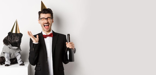 Heureux jeune homme célébrant des vacances avec un chien mignon tenant du champagne et souriant carlin et propriétaire wearin