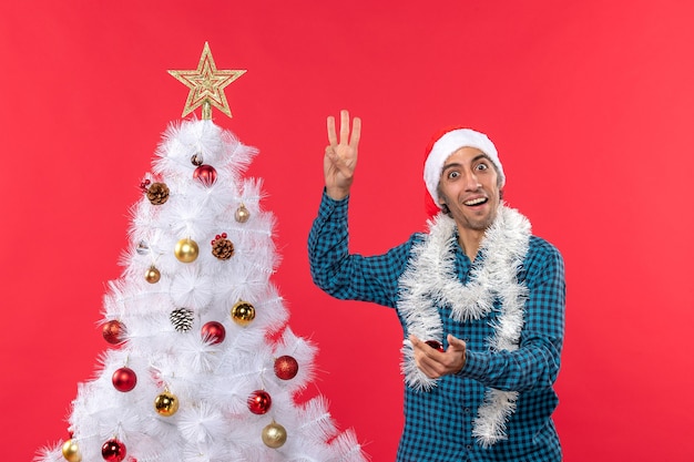 Heureux jeune homme avec chapeau de père Noël dans une chemise dépouillé bleu et montrant trois arbre de Noël sur rouge