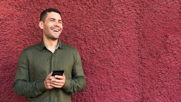 Heureux jeune homme chaume tenant un téléphone portable debout près d&#39;un mur rugueux