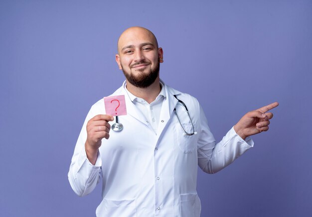 Heureux jeune homme chauve portant une robe médicale et un stéthoscope tenant un point d'interrogation en papier et des points sur le côté