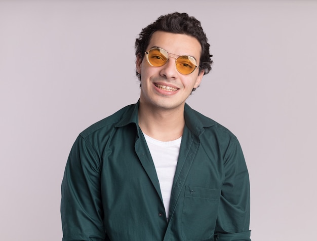 Heureux jeune homme en chemise verte portant des lunettes à l'avant avec le sourire sur le visage debout sur un mur blanc