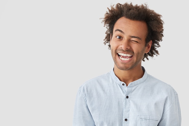 Heureux jeune homme clignote des yeux, a une expression positive, a les cheveux noirs croquants, habillé avec désinvolture