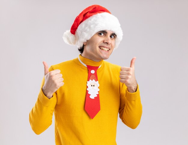 Heureux jeune homme à col roulé jaune et bonnet de noel avec cravate drôle regardant la caméra avec le sourire sur le visage montrant les pouces vers le haut debout sur fond blanc