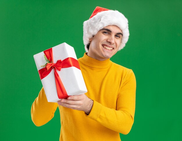 Heureux jeune homme à col roulé jaune et bonnet de Noel tenant un cadeau regardant la caméra en souriant joyeusement debout sur fond vert