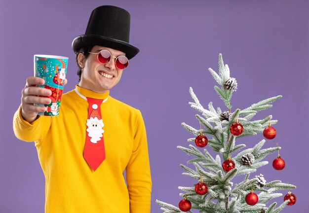 Heureux jeune homme à col roulé jaune et lunettes portant chapeau noir et cravate drôle montrant tasse de papier coloré souriant joyeusement debout à côté d'un arbre de Noël sur fond violet