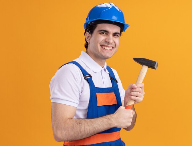 Heureux jeune homme constructeur en uniforme de construction et casque de sécurité tenant un marteau à l'avant souriant joyeusement debout sur un mur orange