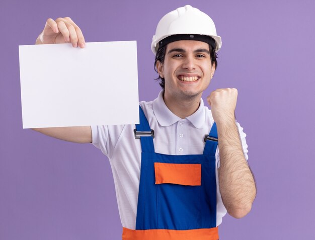 Heureux jeune homme constructeur en uniforme de construction et casque de sécurité tenant une page blanche à l'avant avec le sourire sur le visage serrant le poing debout sur le mur violet