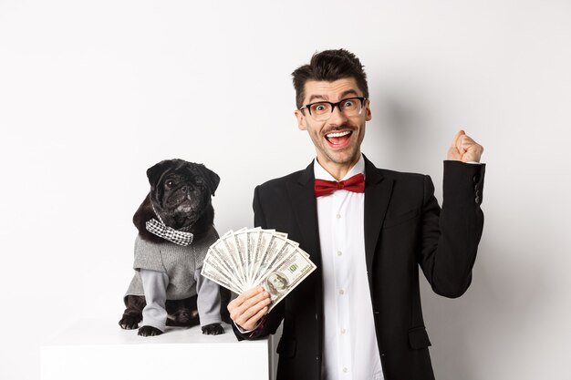 Heureux jeune homme en costume gagne de l'argent avec son chien. Guy se réjouit, tenant des dollars, un carlin noir en costume regardant la caméra, fond blanc.