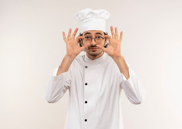 Heureux jeune homme cuisinier portant l'uniforme de chef et des lunettes mettant la main sur des verres isolés sur un mur blanc