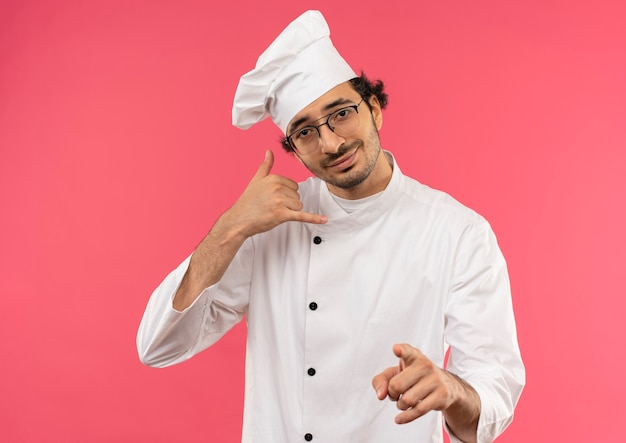 Heureux jeune homme cuisinier portant l'uniforme de chef et des lunettes montrant le geste de téléphone d'appel avec vous geste sur rose