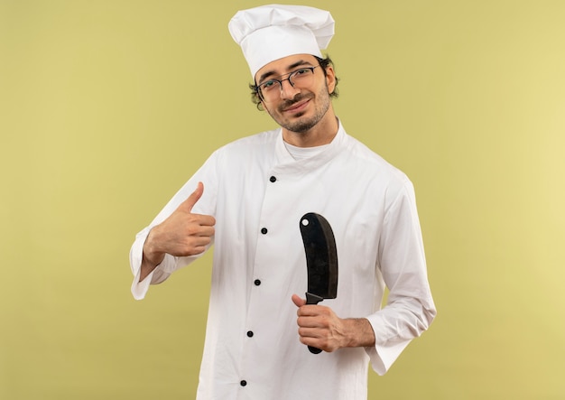 Heureux jeune homme cuisinier portant l'uniforme de chef et des lunettes tenant couperet son pouce vers le haut isolé sur mur vert