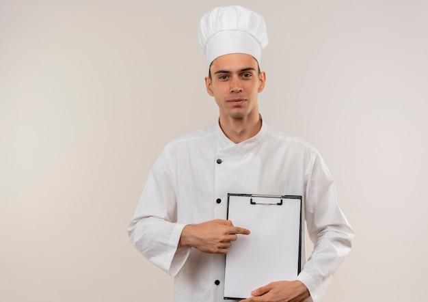 Heureux jeune homme cuisinier portant l'uniforme de chef pointe le doigt sur le presse-papiers dans sa main avec copie espace