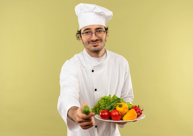 Heureux jeune homme cuisinier portant l'uniforme de chef et des verres tenant des légumes sur une assiette et tenant le concombre isolé sur un mur vert