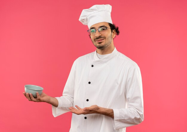 Heureux jeune homme cuisinier portant l'uniforme de chef et verres tenant et points avec la main au bol isolé sur mur rose