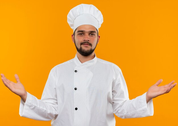 Heureux jeune homme cuisinier en uniforme de chef montrant les mains vides et à la isolé sur l'espace orange