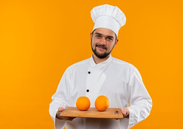 Heureux jeune homme cuisinier en uniforme de chef tenant une planche à découper avec des oranges sur elle à isolé sur l'espace orange