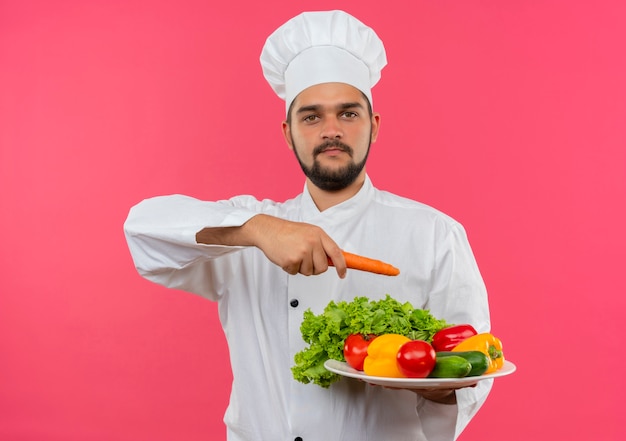 Heureux jeune homme cuisinier en uniforme de chef tenant la plaque de légumes et pointant avec la carotte à elle isolé sur l'espace rose