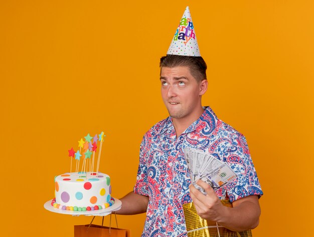 Heureux jeune homme de fête portant une casquette d'anniversaire tenant des cadeaux avec un gâteau et de l'argent isolé sur orange