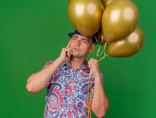 Heureux jeune homme de fête portant un chapeau bleu tenant des ballons et mettant la main sur la joue isolé sur vert