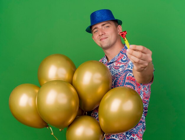 Heureux jeune homme de fête portant un chapeau bleu tenant des ballons et tenant un ventilateur de fête isolé sur vert