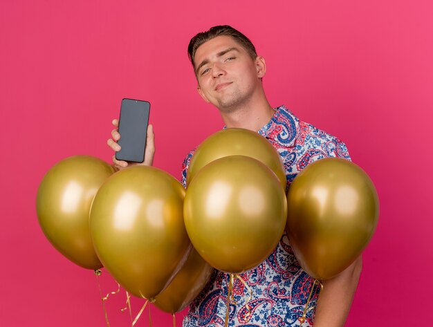 Heureux jeune homme de fête portant une chemise colorée debout parmi les ballons et tenant le téléphone isolé sur rose