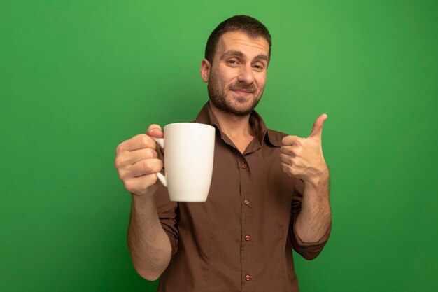 Heureux jeune homme isolé sur vert tenant une tasse de thé montrant le pouce vers le haut du mur