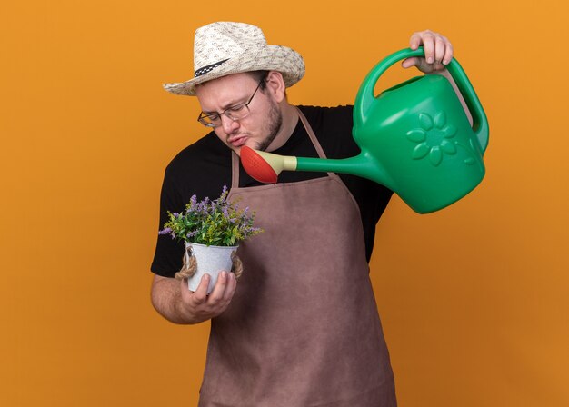 Heureux jeune homme jardinier portant chapeau de jardinage et gants d'arrosage fleur en pot de fleurs avec arrosoir isolé sur mur orange