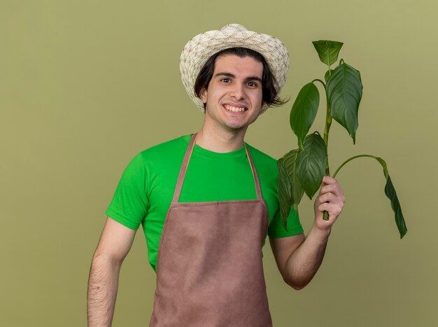 Heureux jeune homme jardinier en tablier et hat holding plant regardant la caméra avec le sourire sur le visage debout sur fond clair