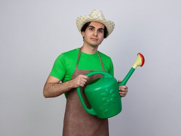 Heureux jeune homme jardinier en uniforme portant un chapeau de jardinage tenant un arrosoir