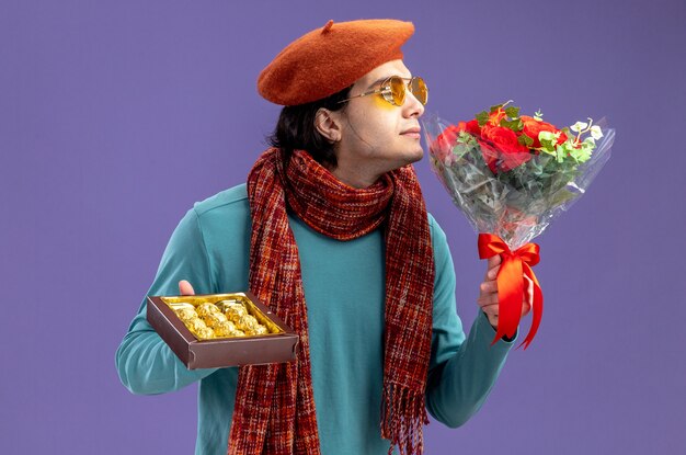 Heureux jeune homme le jour de la Saint-Valentin portant un chapeau avec une écharpe et des lunettes tenant une boîte de bonbons reniflant le bouquet dans sa main isolé sur fond bleu