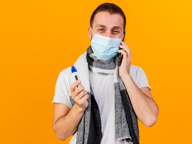 Heureux jeune homme malade portant un chapeau d'hiver et un masque médical parle au téléphone et tenant un thermomètre isolé sur fond jaune