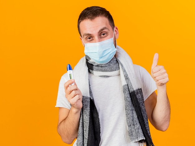 Heureux jeune homme malade portant un chapeau d'hiver et un masque médical tenant un thermomètre montrant le pouce vers le haut isolé sur fond jaune