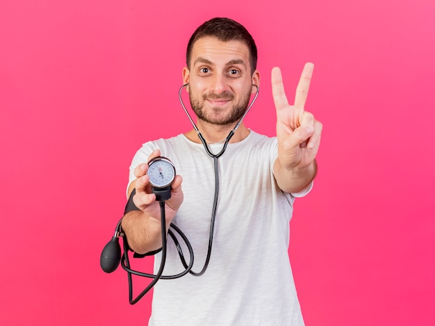 Heureux jeune homme malade portant un stéthoscope tenant un sphygmomanomètre montrant le geste de paix isolé sur fond rose