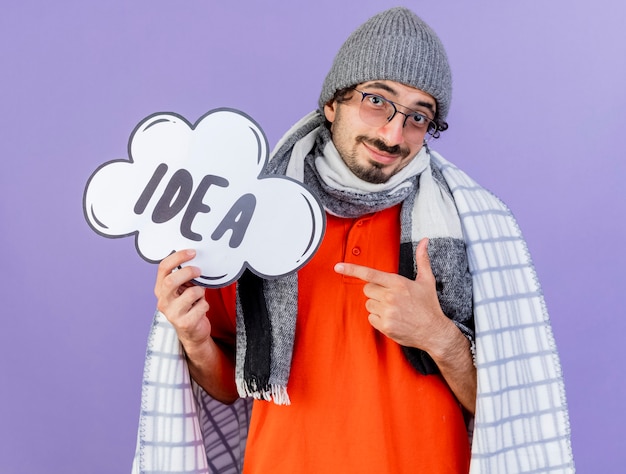 Heureux jeune homme malade de race blanche portant des lunettes chapeau d'hiver et écharpe enveloppé dans un plaid tenant et pointant sur la bulle d'idée regardant la caméra isolée sur fond violet