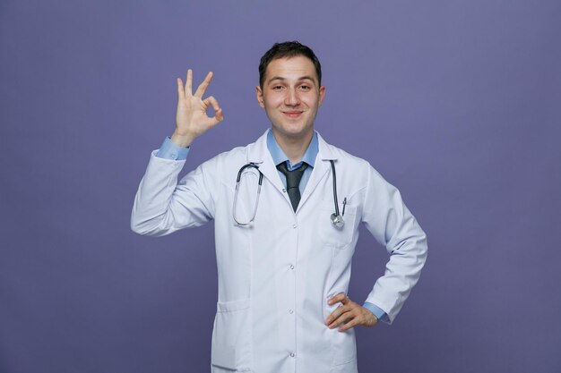 Heureux jeune homme médecin portant une robe médicale et un stéthoscope autour du cou en gardant la main sur la taille en regardant la caméra montrant le signe ok isolé sur fond violet