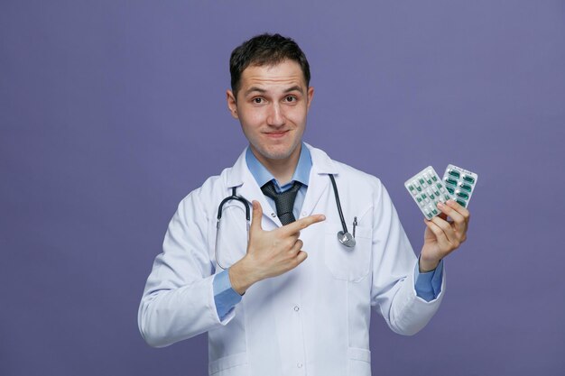 Heureux jeune homme médecin portant une robe médicale et un stéthoscope autour du cou regardant la caméra montrant des paquets de pilules pointant vers eux isolés sur fond violet