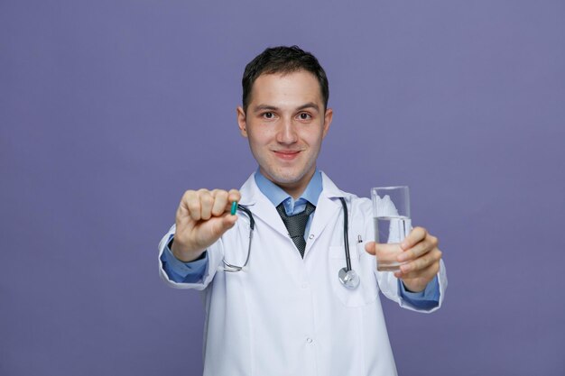 Heureux jeune homme médecin portant une robe médicale et un stéthoscope autour du cou regardant la caméra qui s'étend de la capsule et du verre d'eau vers la caméra isolée sur fond violet