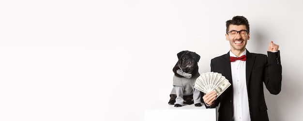 Photo gratuite heureux jeune homme et mignon chien noir debout dans des costumes de fête propriétaire de carlin détenant de l'argent dollars et re