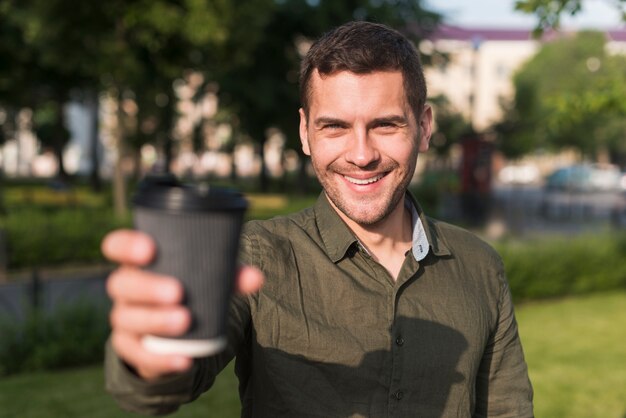 Heureux jeune homme montrant une tasse de café jetable au parc