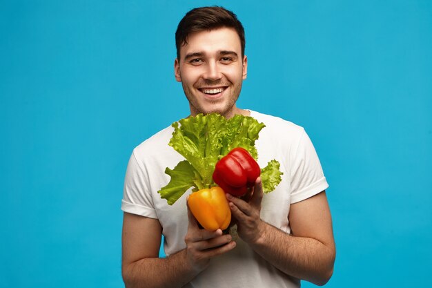 Heureux jeune homme non rasé végétalien avec un corps musclé avec un large sourire radieux transportant des légumes colorés frais et de la laitue de l'épicerie. Veganisme, nourriture crue et régime