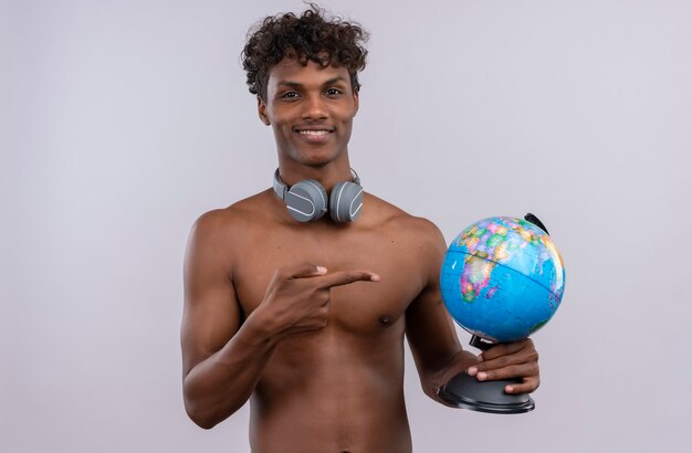 Un heureux jeune homme à la peau foncée beau avec des cheveux bouclés portant des écouteurs pointant sur globe avec index