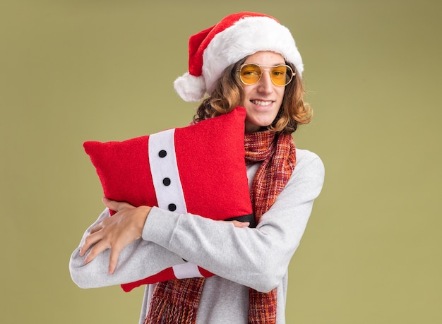 Heureux jeune homme portant un bonnet de noel et des lunettes jaunes avec un foulard chaud autour du cou tenant un oreiller de noël avec un sourire sur le visage debout sur un mur vert