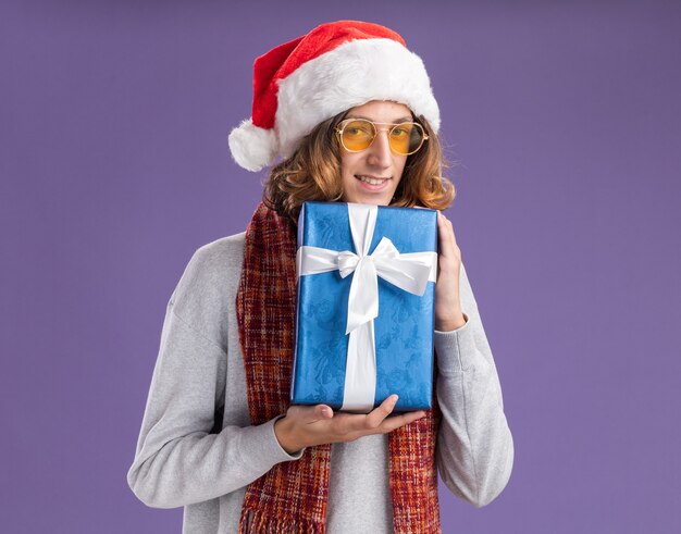 Heureux jeune homme portant un bonnet de noel de Noël et des lunettes jaunes avec un foulard chaud autour de son cou tenant le cadeau de Noël regardant la caméra en souriant joyeusement debout sur fond violet